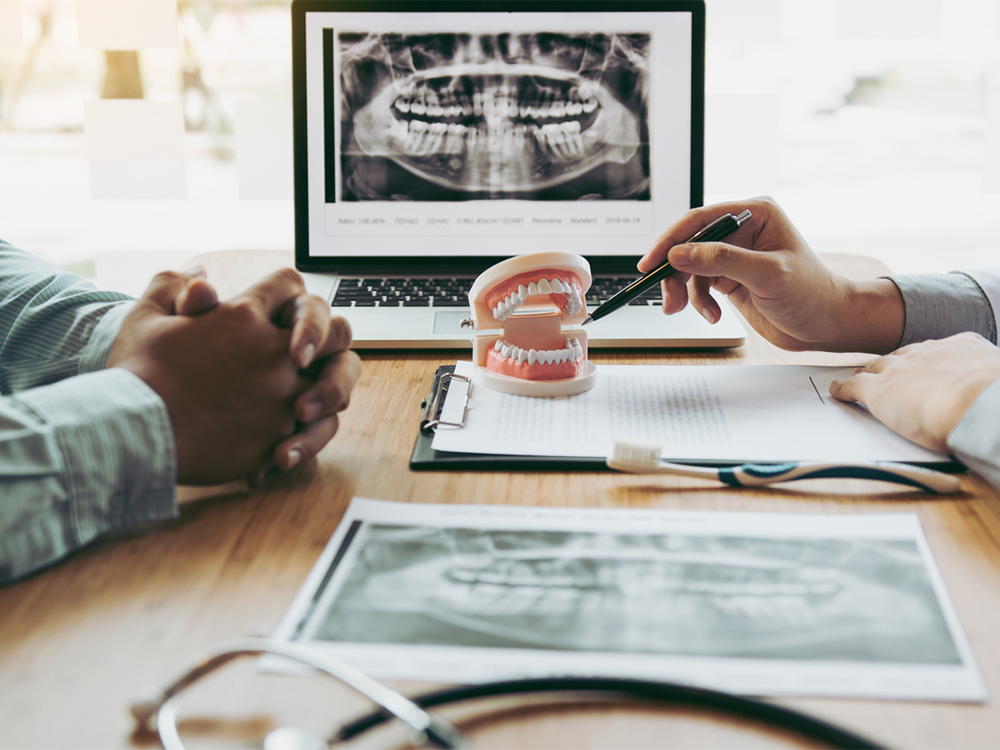 a desk with x-rays on a computer and a dentist pointing to a jaw model with a notepad in front of him