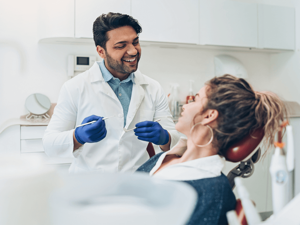A male dentist in a white coat and gloves talks with a smiling patient in a bright, modern dental office.
