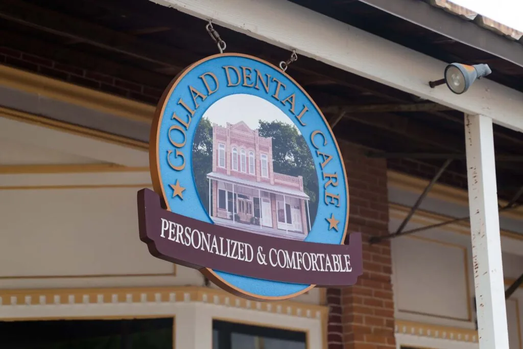 A hanging wooden sign for "Goliad Dental Care" with the tagline "Personalized & Comfortable," featuring an illustration of a historic building.