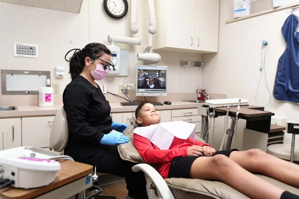 A dental assistant in black scrubs and gloves gently prepares a young patient in a red shirt for a procedure in a modern treatment room.