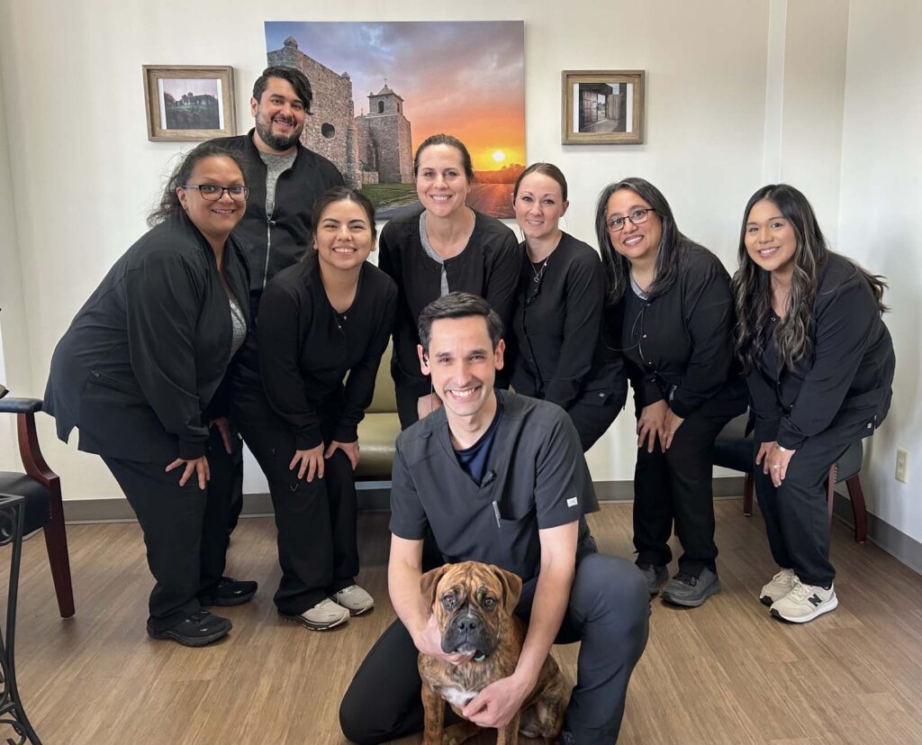 The Goliad Dental Care team, dressed in black scrubs, poses together for a group photo in the office, with Bella the therapy dog sitting in front.