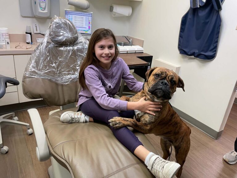 A young girl in a purple outfit sits in a dental chair, happily hugging Bella, the therapy dog, providing comfort in the dental office.