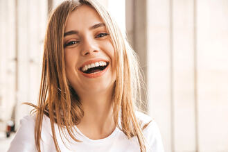 A smiling person with long light brown hair wearing a white shirt, posed in front of softly lit vertical window panels.