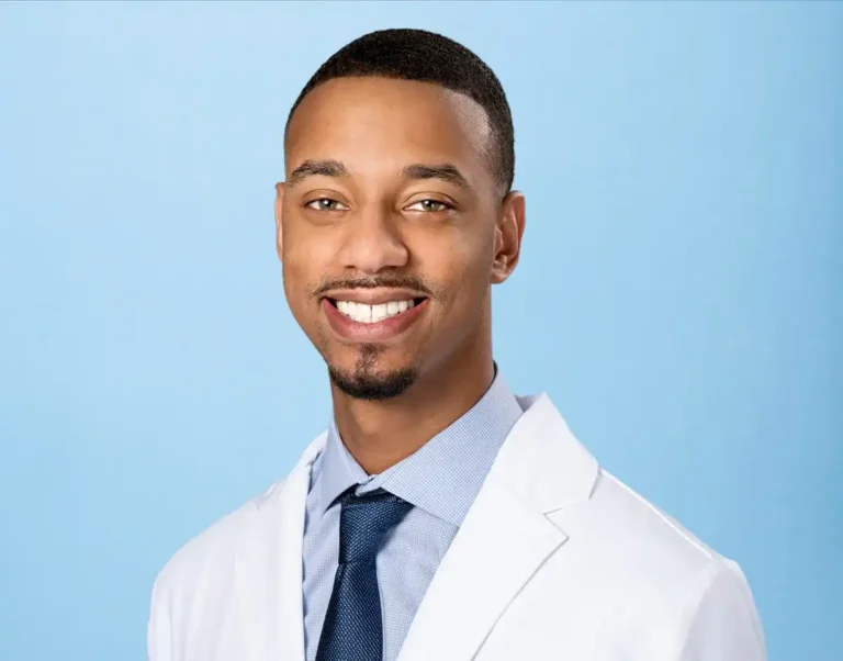 Professional portrait of Dr. Williams in white lab coat, light blue shirt, and dark blue tie, smiling against solid light blue background.